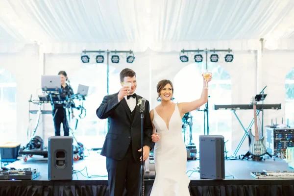 capa post: Noivos em casamento brindando juntos no palco, com microfone e banda ao fundo em uma tenda decorada.