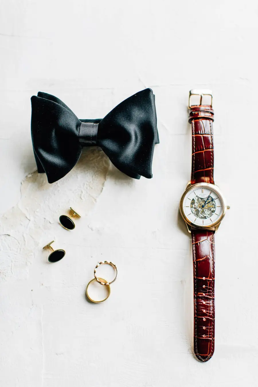 Conjunto de acessórios masculinos para casamento sobre superfície clara, incluindo gravata borboleta preta, relógio com pulseira de couro marrom, abotoaduras pretas e douradas e duas alianças douradas.