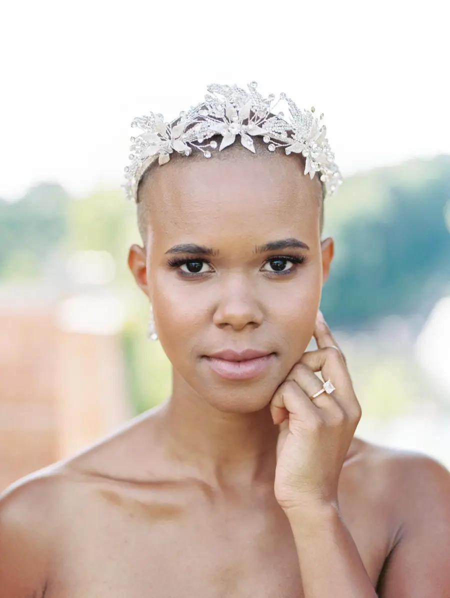 Noiva com cabeça raspada usando tiara prateada com detalhes florais e brincos delicados, tocando o rosto com a mão