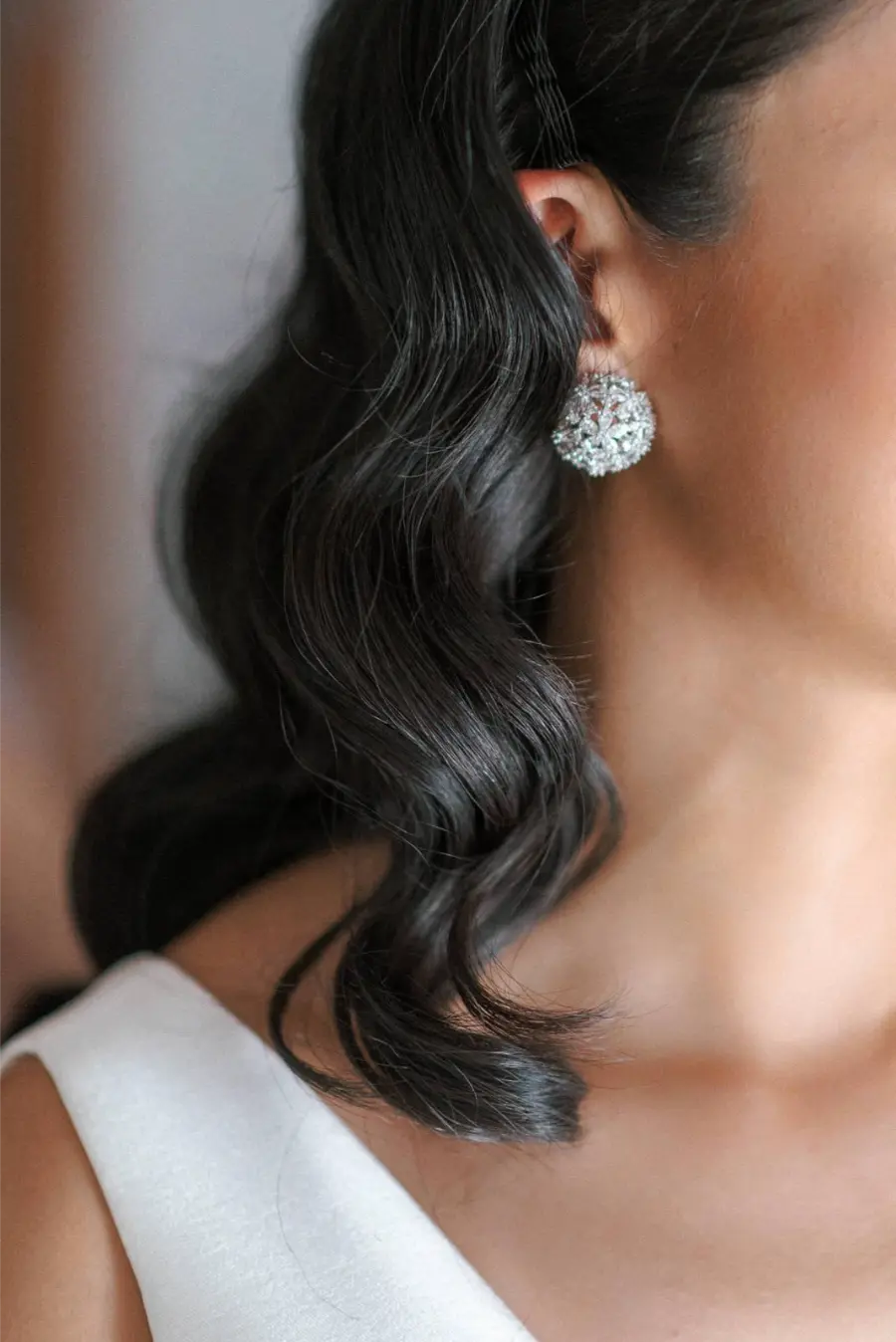 Close no rosto e cabelo castanho ondulado de noiva, mostrando brinco redondo prateado com pedras brilhantes.