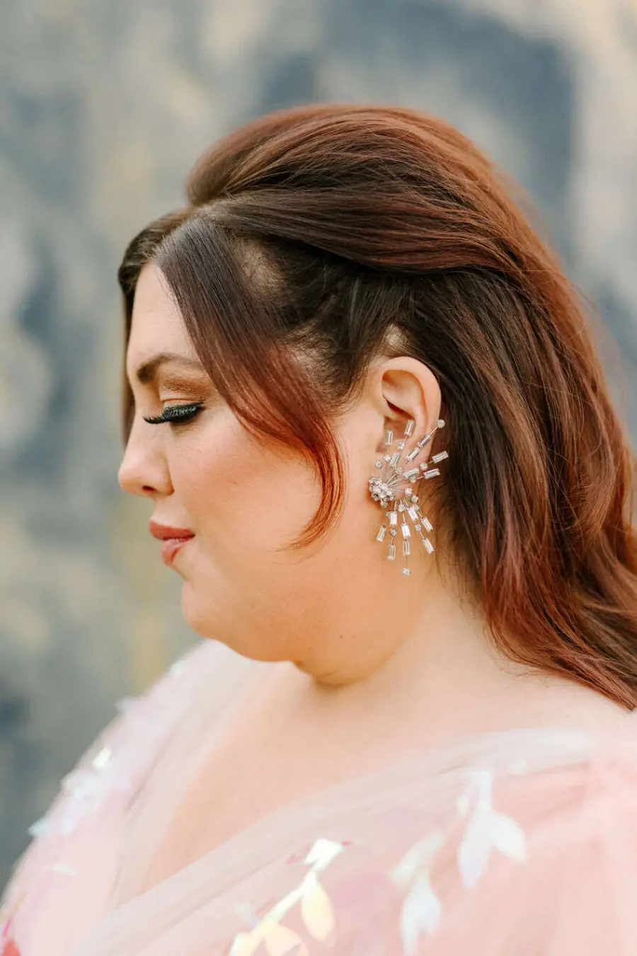 Noiva de perfil com cabelo castanho-avermelhado semi-preso, usando brinco grande prateado em formato geométrico e maquiagem suave