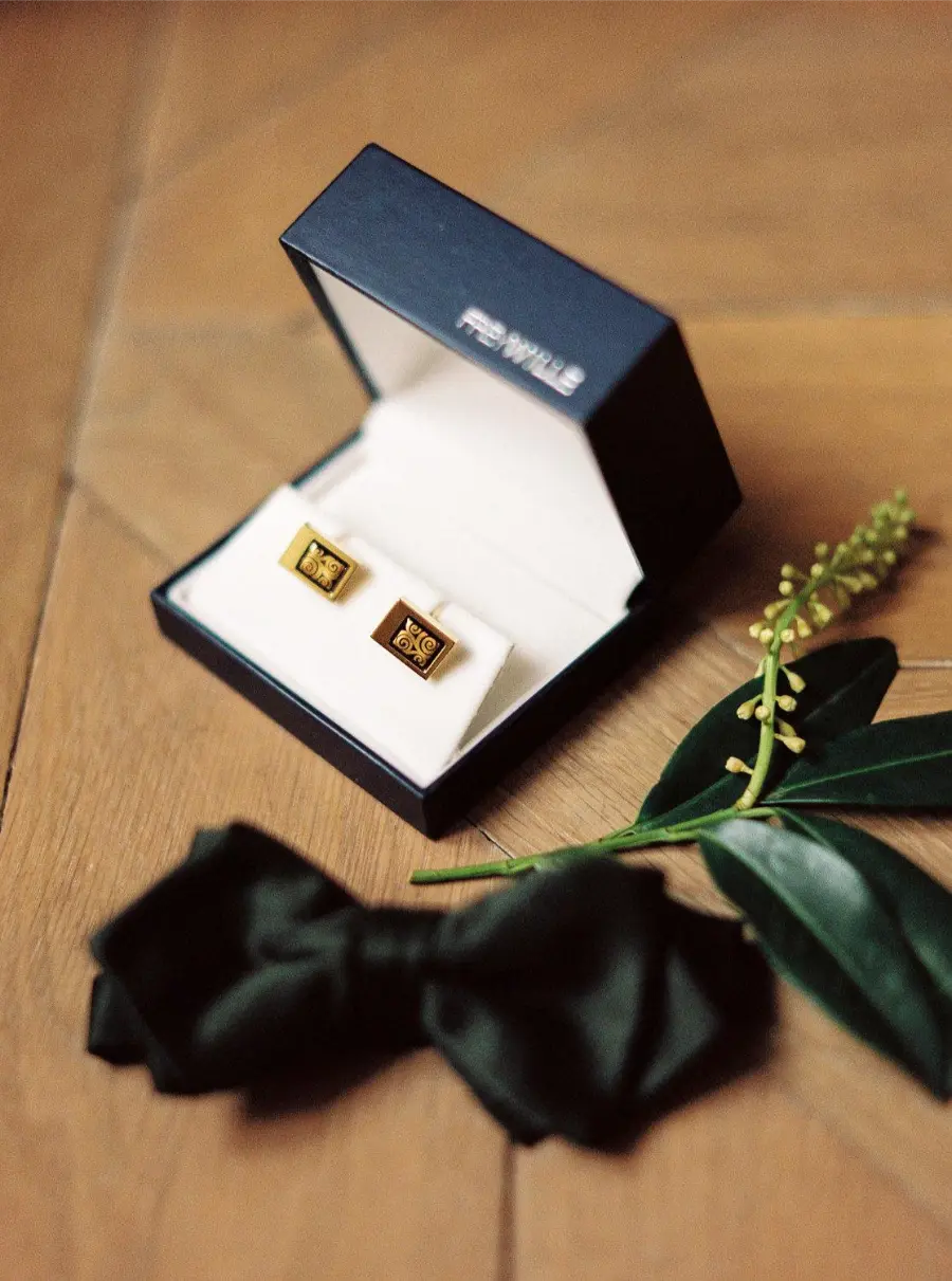 Caixa aberta com par de abotoaduras douradas sobre base branca, ao lado de gravata borboleta preta e ramo de folhas sobre piso de madeira