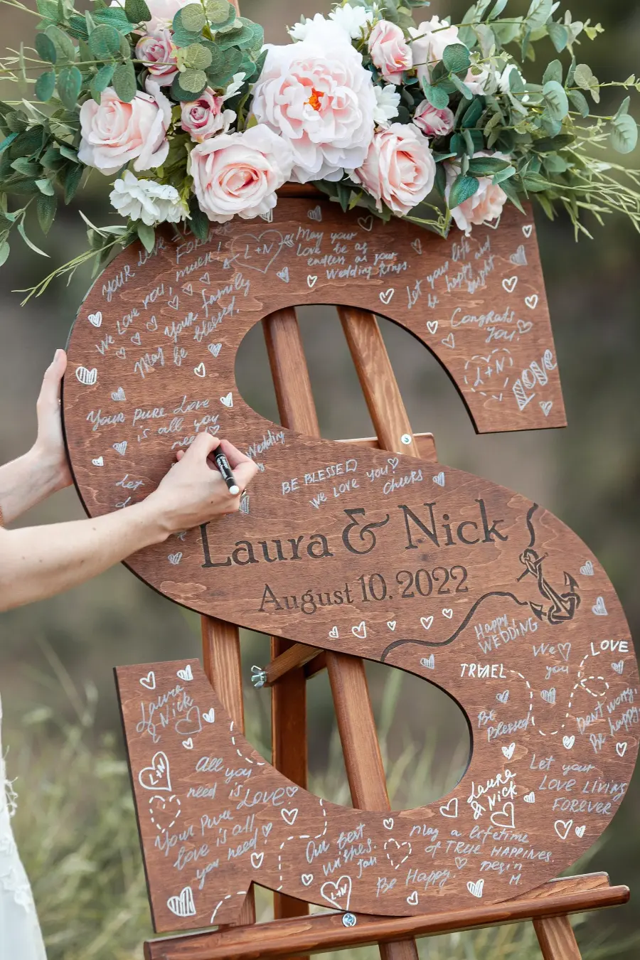 Placa de madeira em formato da letra S com mensagens escritas por convidados em casamento decorada com flores.