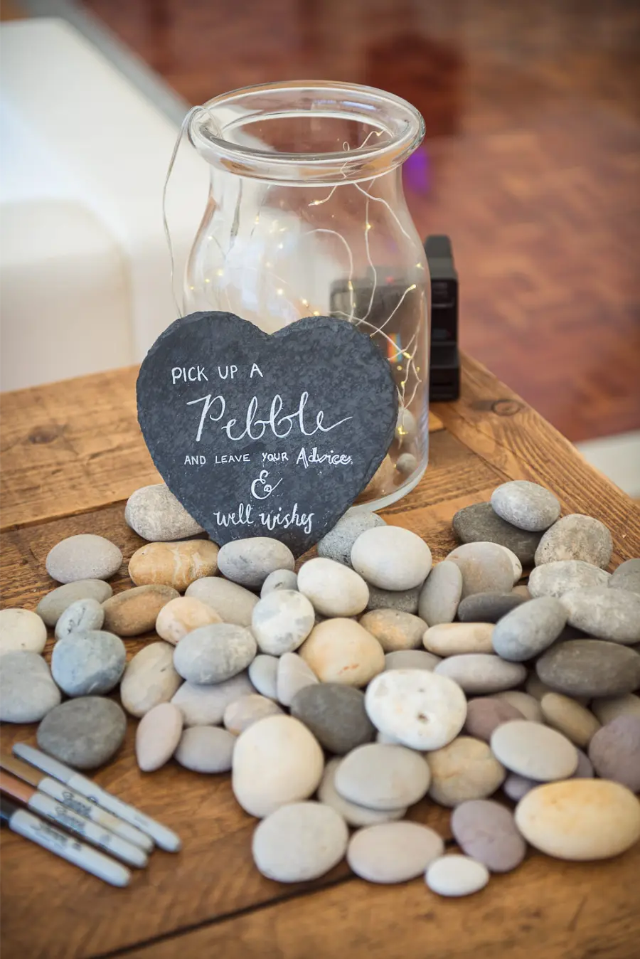 Um pote de vidro transparente ao lado de várias pedras lisas, acompanhado de um coração de ardósia com instrução para que os convidados escrevam mensagens e conselhos nos pedrinhas.