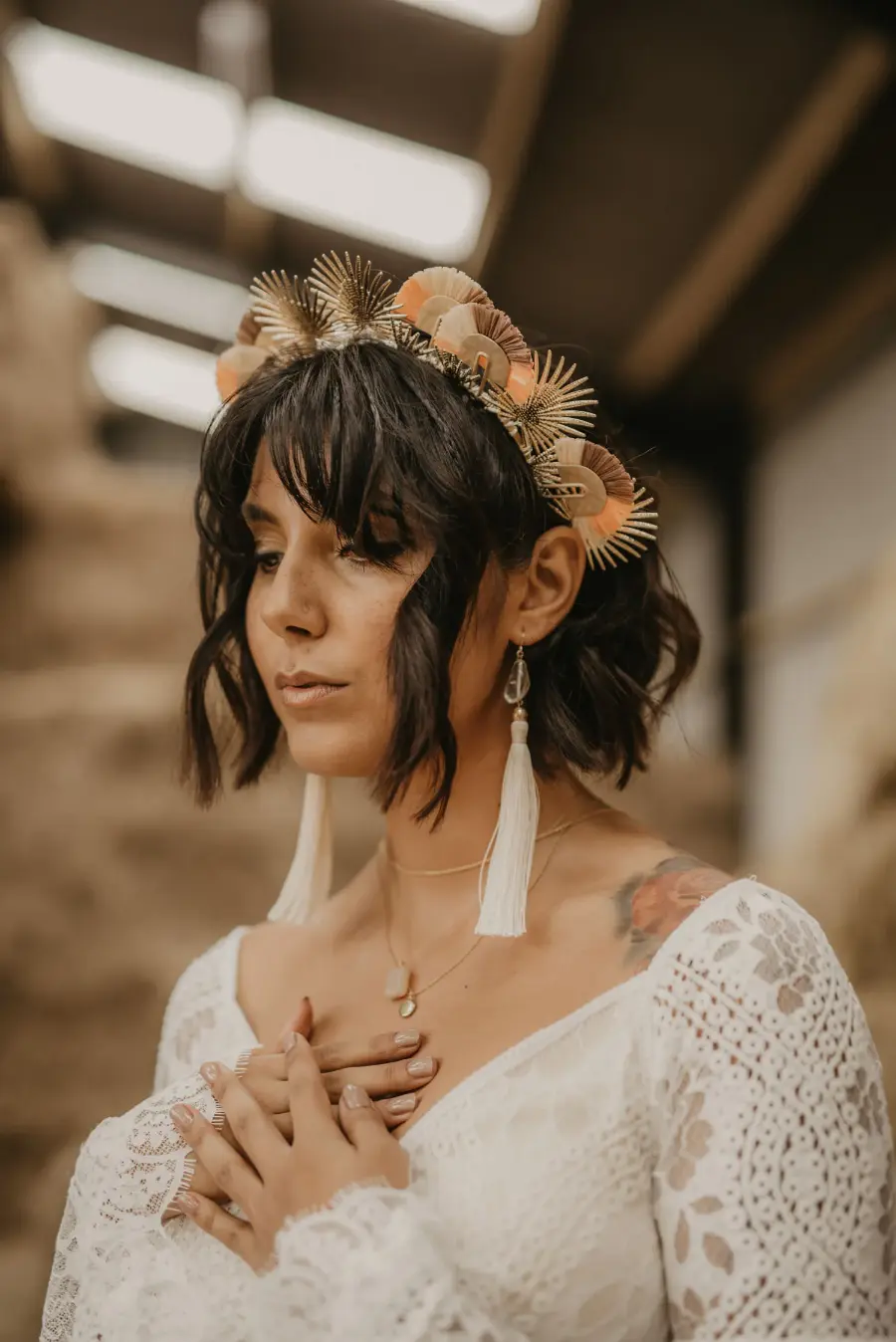 Noiva com vestido de renda branca, cabelos curtos e coroa de flores e elementos dourados, usando brincos longos de tassel branco