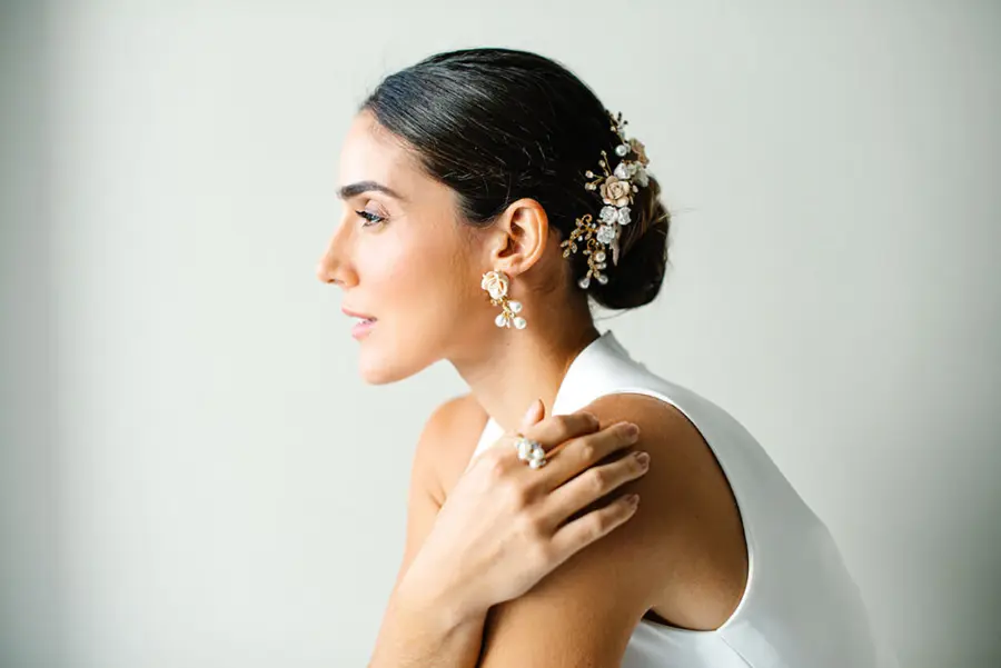 Noiva de perfil com penteado preso adornado com arranjo floral dourado e pérolas, usando brincos e anel combinando, vestindo vestido branco sem mangas