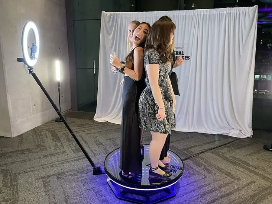 Grupo de amigas sorrindo e posando juntas em uma plataforma 360 iluminada, durante evento noturno com fundo personalizado.
