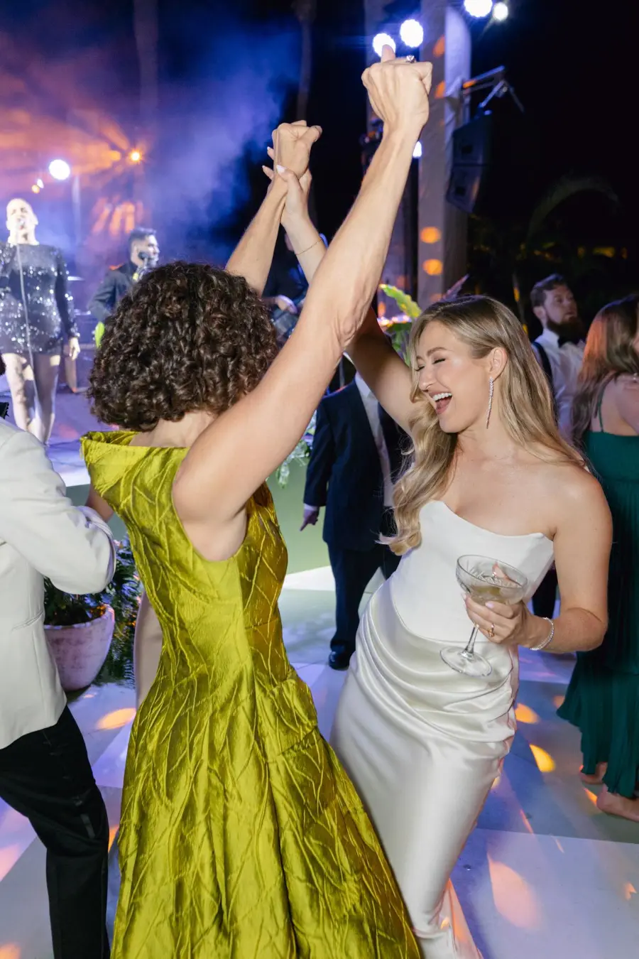 Convidada e noiva dan&ccedil;ando animados na pista de casamento com ilumina&ccedil;&atilde;o de palco e banda ao vivo ao fundo.