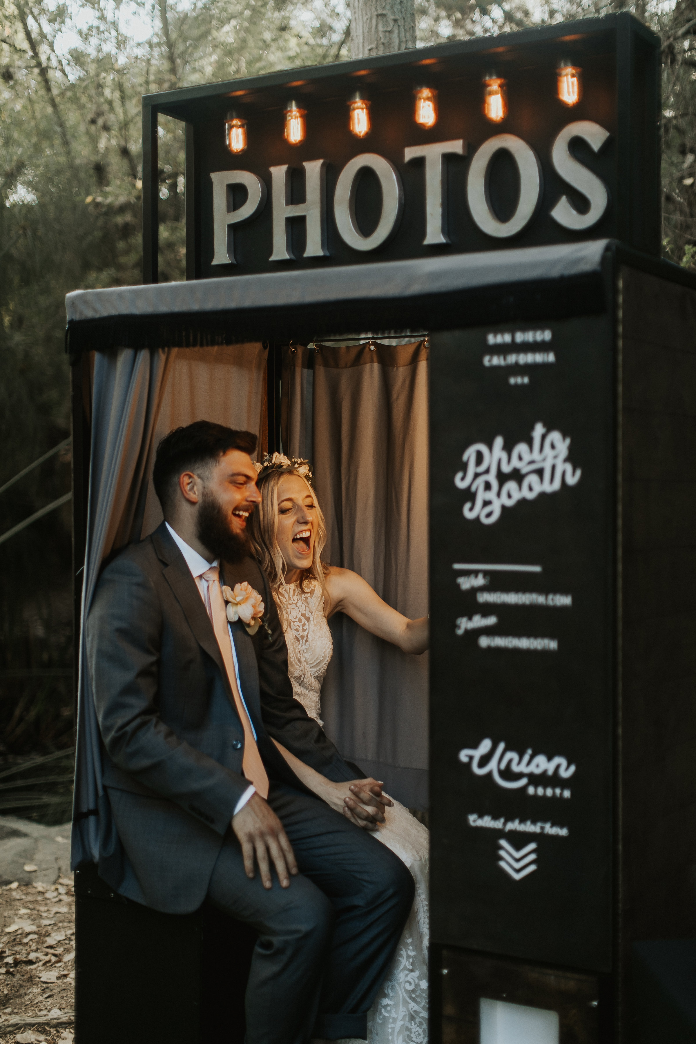 Noivos se divertindo dentro de uma cabine de fotos de casamento, sorrindo enquanto posam juntos.