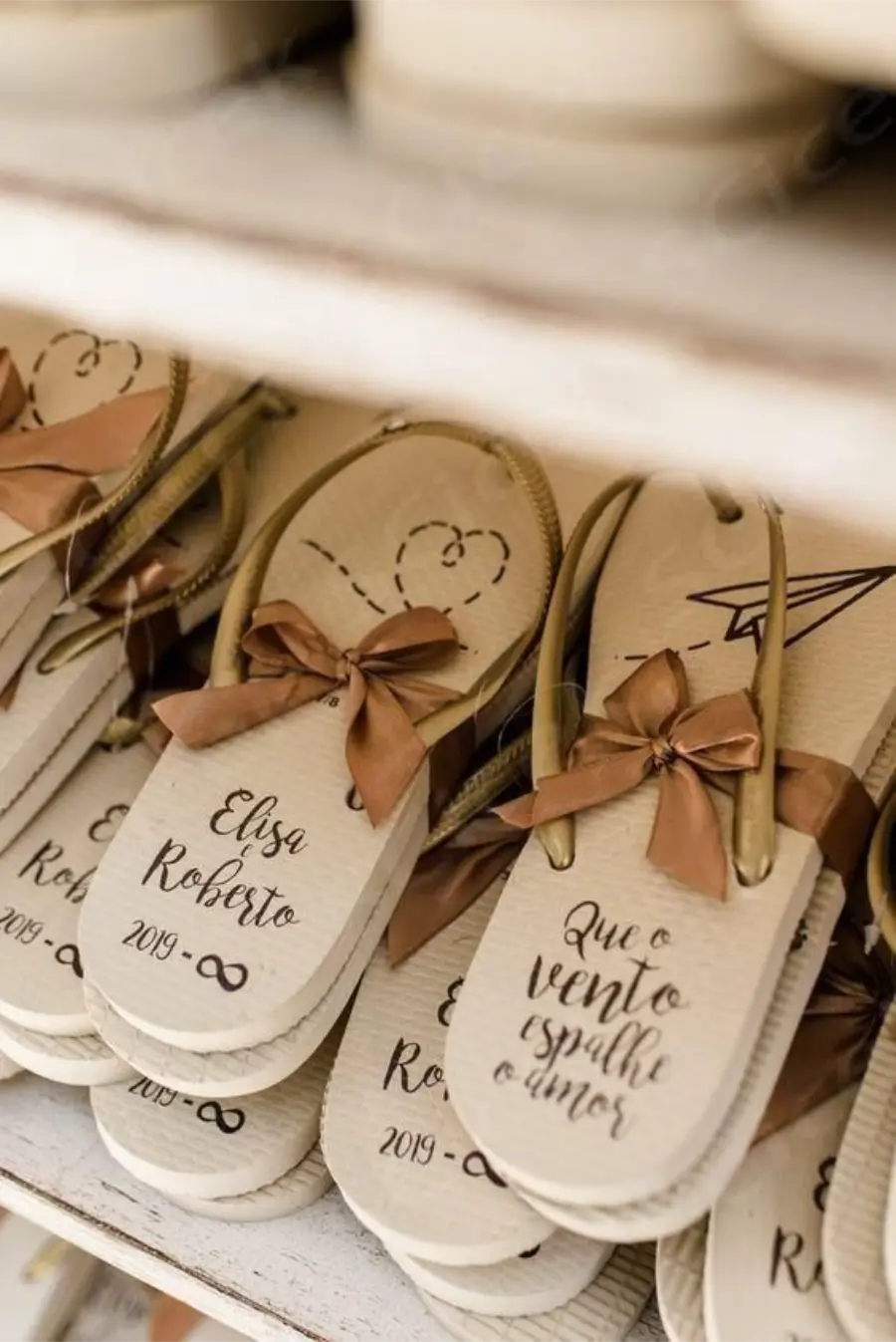 Chinelos personalizados de casamento com nomes Elisa e Roberto, tiras douradas e laço marrom, com frase romântica impressa no solado.
