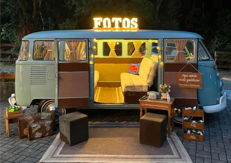 Kombi retrô decorada como cabine de fotos para casamento, iluminada com letreiro “FOTOS” no topo e mobiliário vintage ao redor.