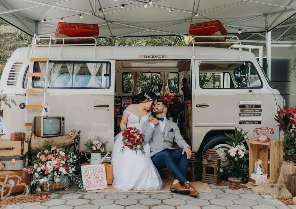 Kombi retrô decorada como cabine de fotos de casamento, com noivos posando em frente à estrutura enfeitada com flores e objetos vintage.