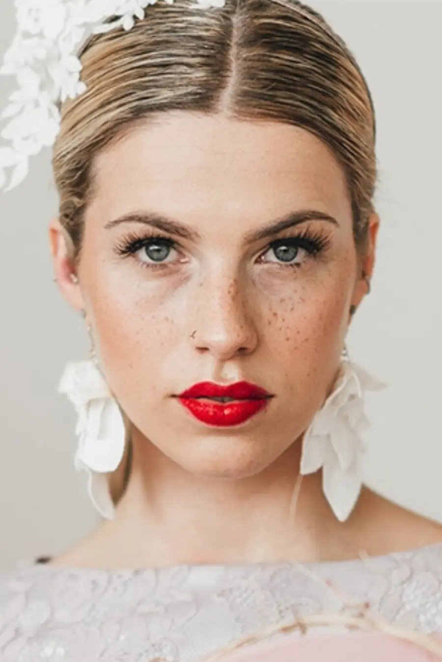 Mulher de cabelo preso e maquiagem marcante com batom vermelho, usando brincos grandes brancos em formato de flores e acessório de cabelo branco.