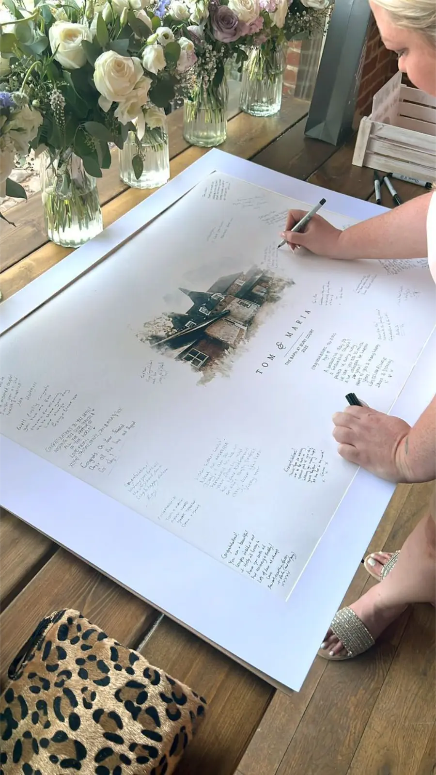 Convidada escrevendo mensagem em quadro de assinatura personalizado com ilustração da fachada de uma casa e nomes dos noivos.
