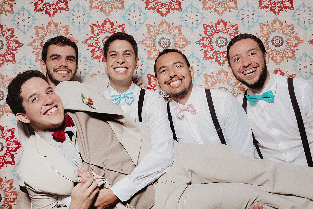 Grupo de amigos com trajes formais posando de forma divertida diante de um painel colorido em cabine de fotos de casamento.