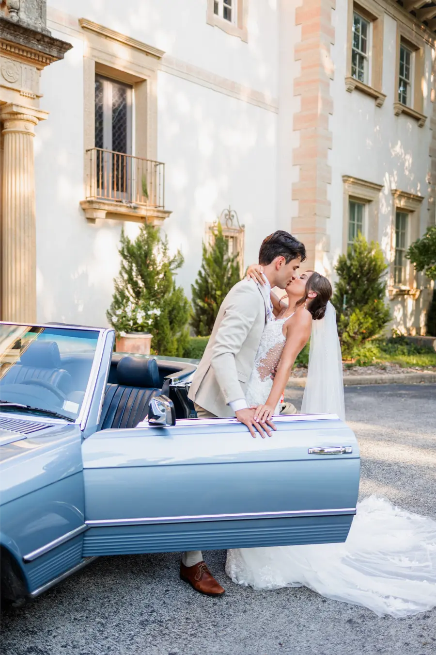 Noivos se beijando ao lado de carro conversível azul claro estacionado em frente a casarão clássico.
