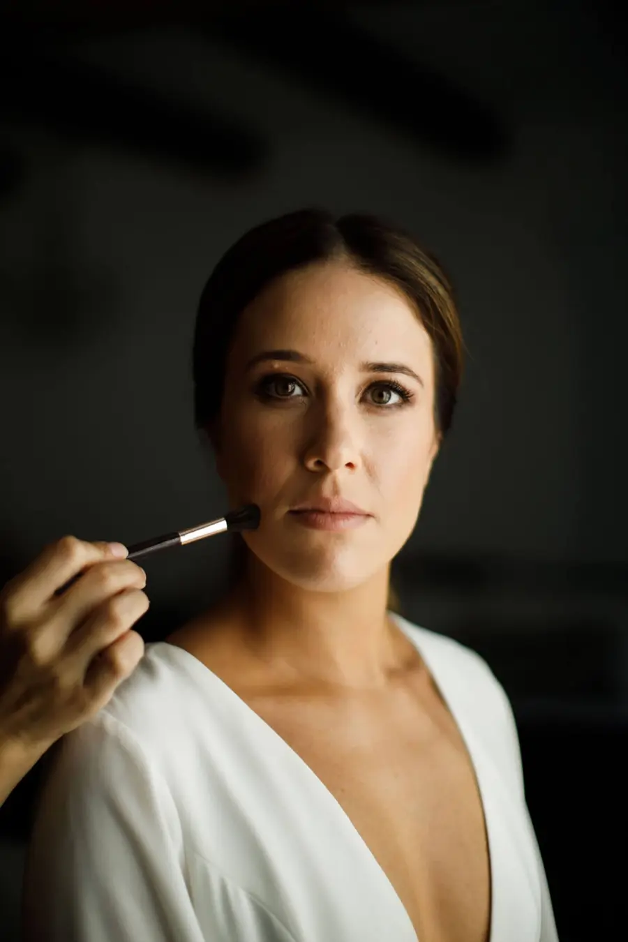 Noiva sendo maquiada, com olhar sereno, recebendo aplicação de blush no rosto