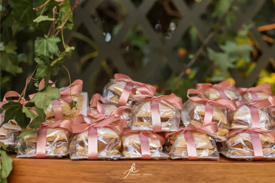 Bem-casados embalados em plástico transparente, com laço rosa, dispostos sobre mesa de madeira como lembrancinhas de casamento.