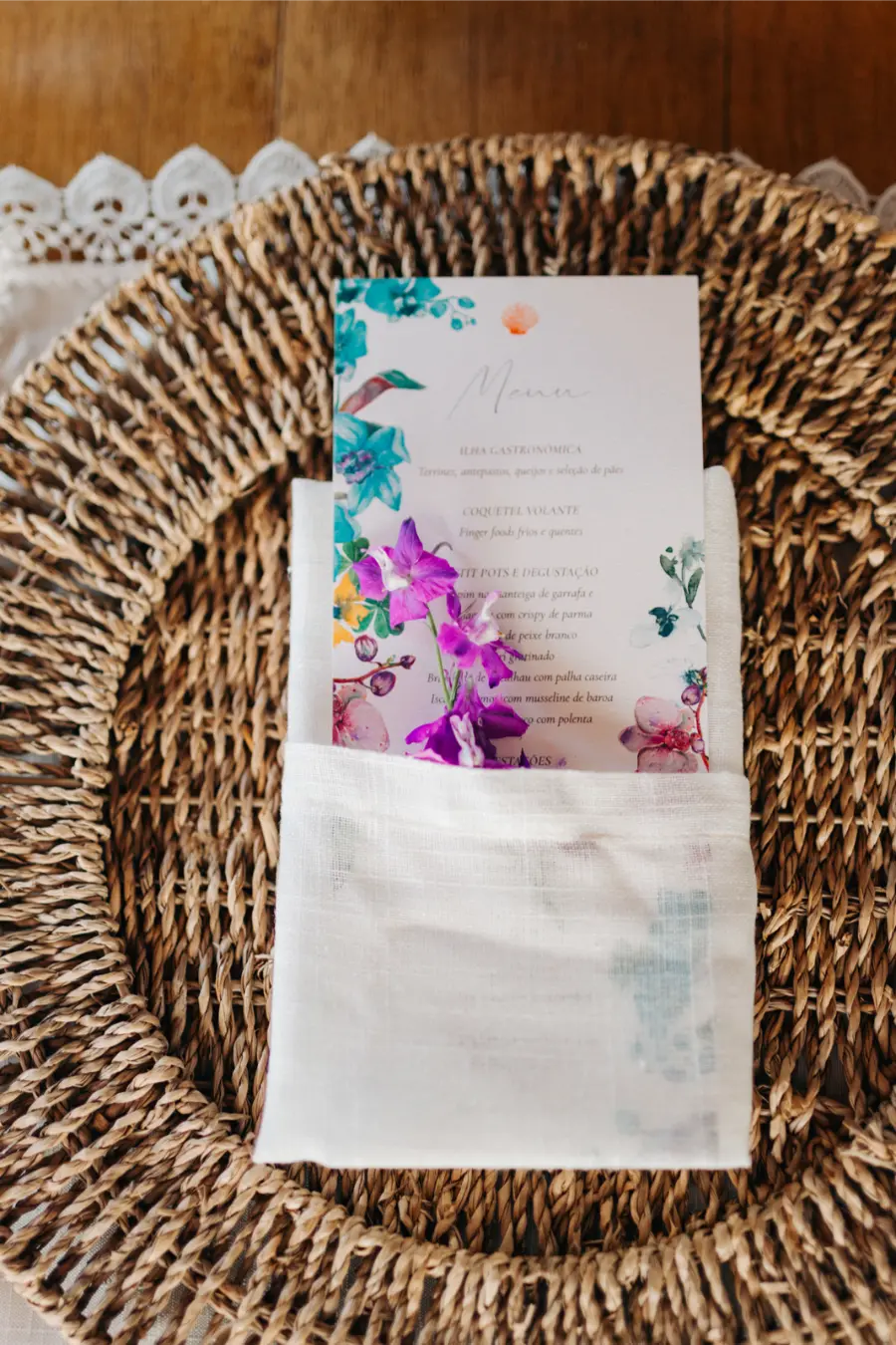 Menu de casamento com ilustração floral em tons vibrantes, disposto em guardanapo branco sobre sousplat de palha.