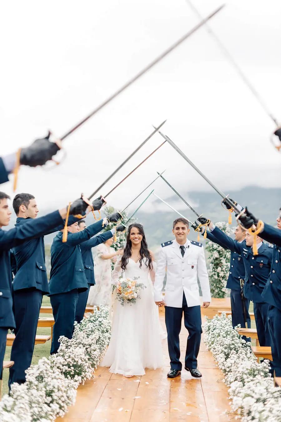Noivos caminham sob t&uacute;nel de espadas formado por militares em farda azul durante cerim&ocirc;nia ao ar livre.