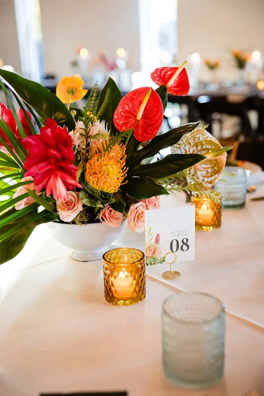 Arranjo floral de mesa de casamento com flores tropicais vibrantes em vermelho, laranja e rosa, acompanhado de velas decorativas e número da mesa.