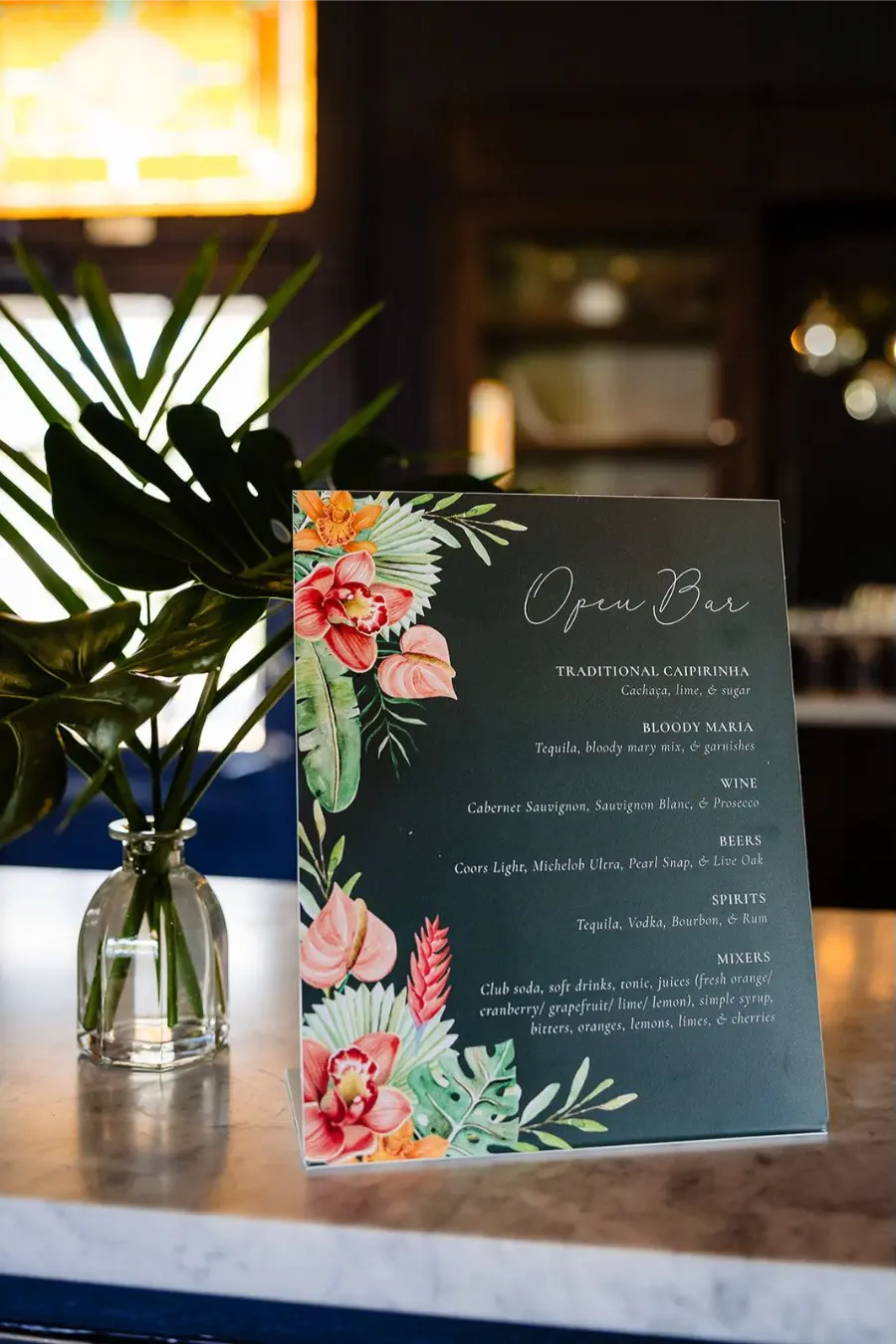 Placa de open bar em casamento com fundo verde escuro, ilustrações tropicais e garrafa decorativa ao lado.