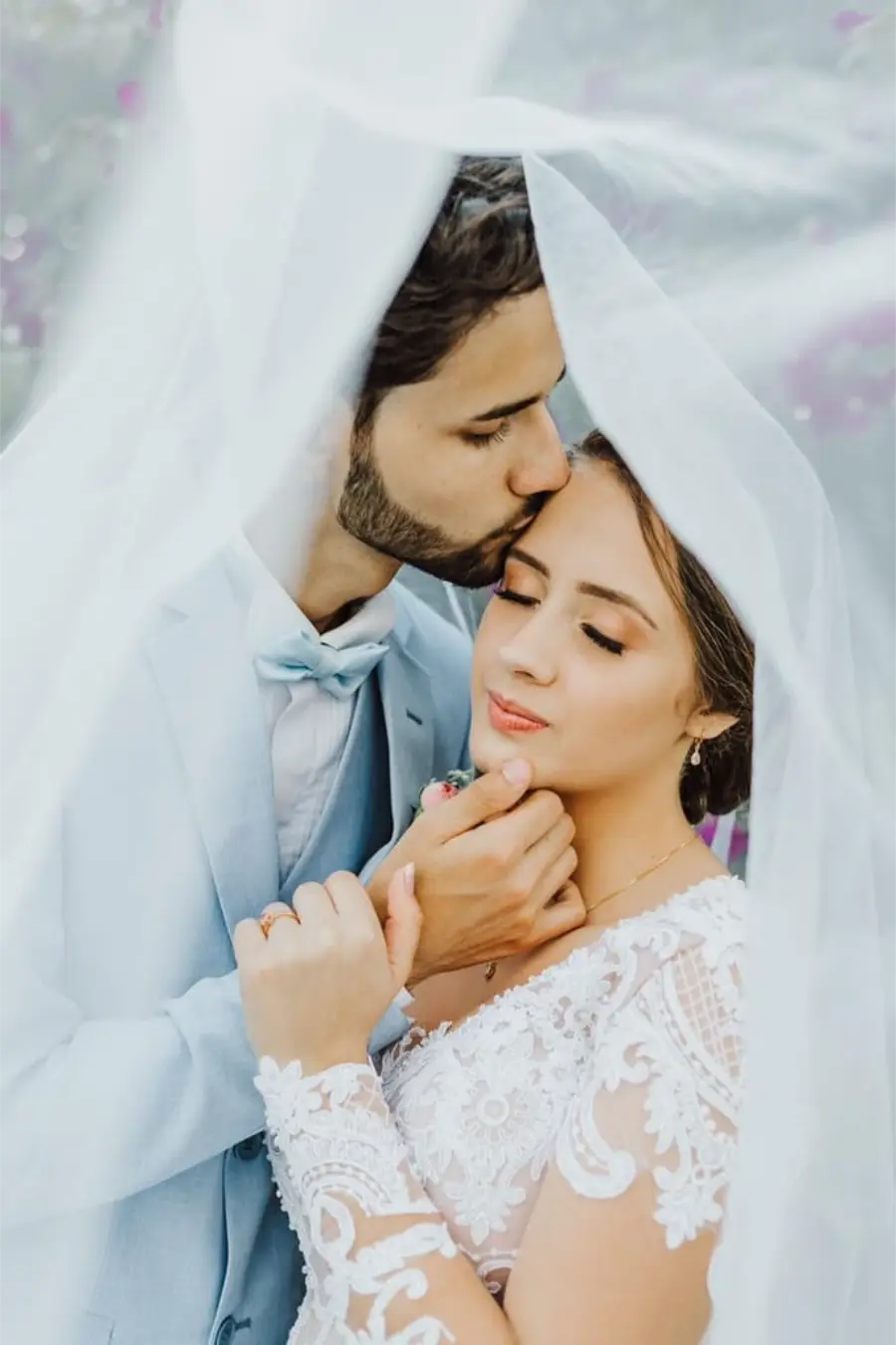Casal de noivos em momento rom&acirc;ntico sob v&eacute;u, noivo com terno azul serenity e noiva com vestido rendado.
