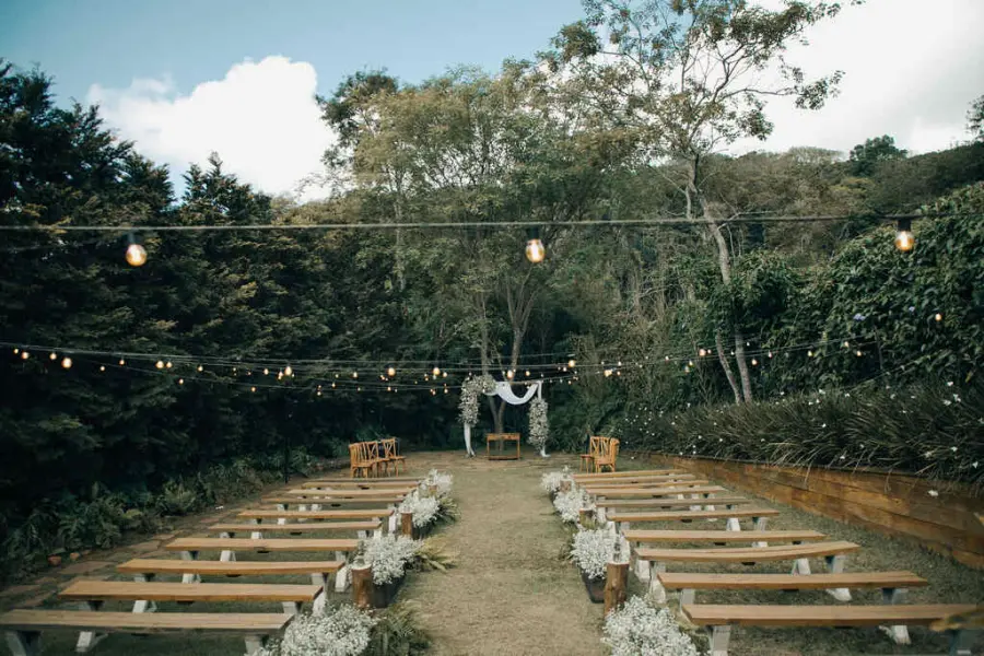 Cerim&ocirc;nia ao ar livre em espa&ccedil;o para casamento r&uacute;stico com bancos de madeira e varal de l&acirc;mpadas