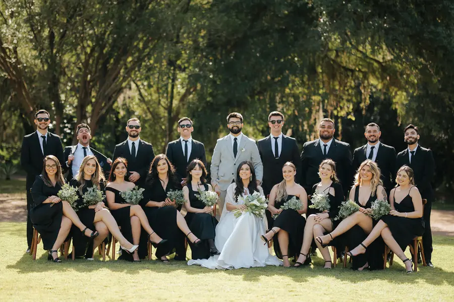 Foto do casal de noivos com seus padrinhos e madrinhas, homens de terno preto e mulheres de vestidos pretos, em ambiente externo.