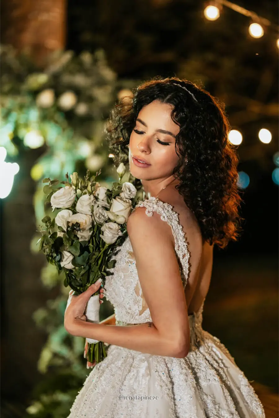 Noiva de cabelos cacheados com vestido rendado e buquê de rosas brancas, posando em cerimônia noturna iluminada por luzes decorativas.