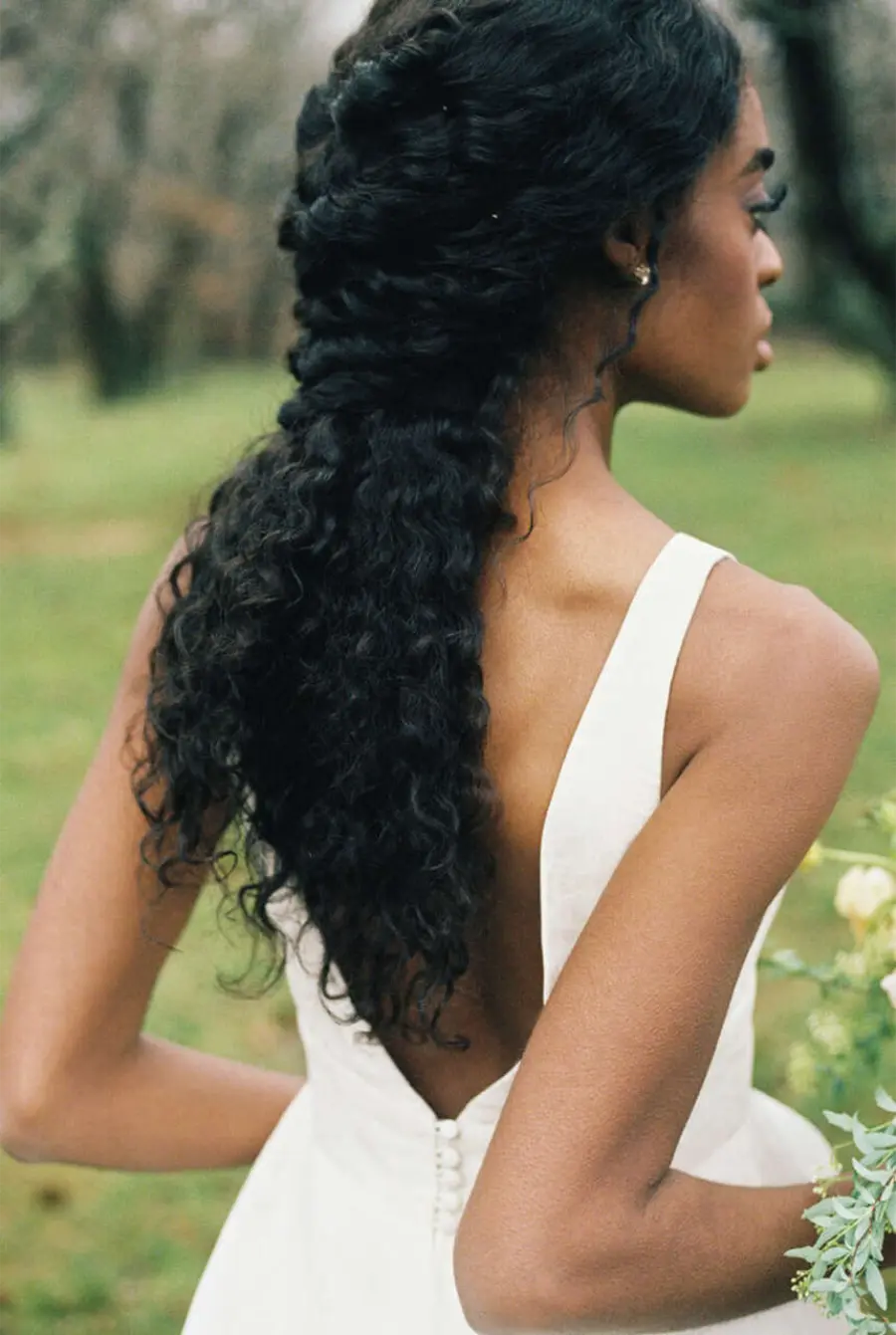 Noiva de costas exibindo penteado solto com trança em cabelo cacheado longo, usando vestido branco com decote profundo nas costas.