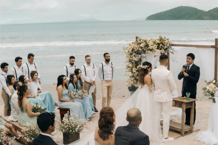 Cerimônia de casamento simples na praia com noivos diante do celebrante e convidados sentados em bancos decorados com flores.