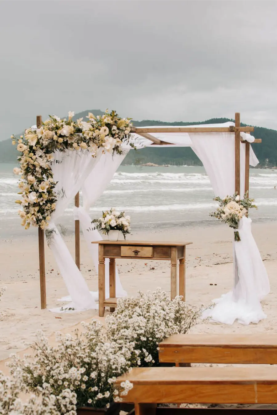 Estrutura de madeira coberta por tecidos brancos e flores claras em frente ao mar, formando cenário para casamento simples na praia.