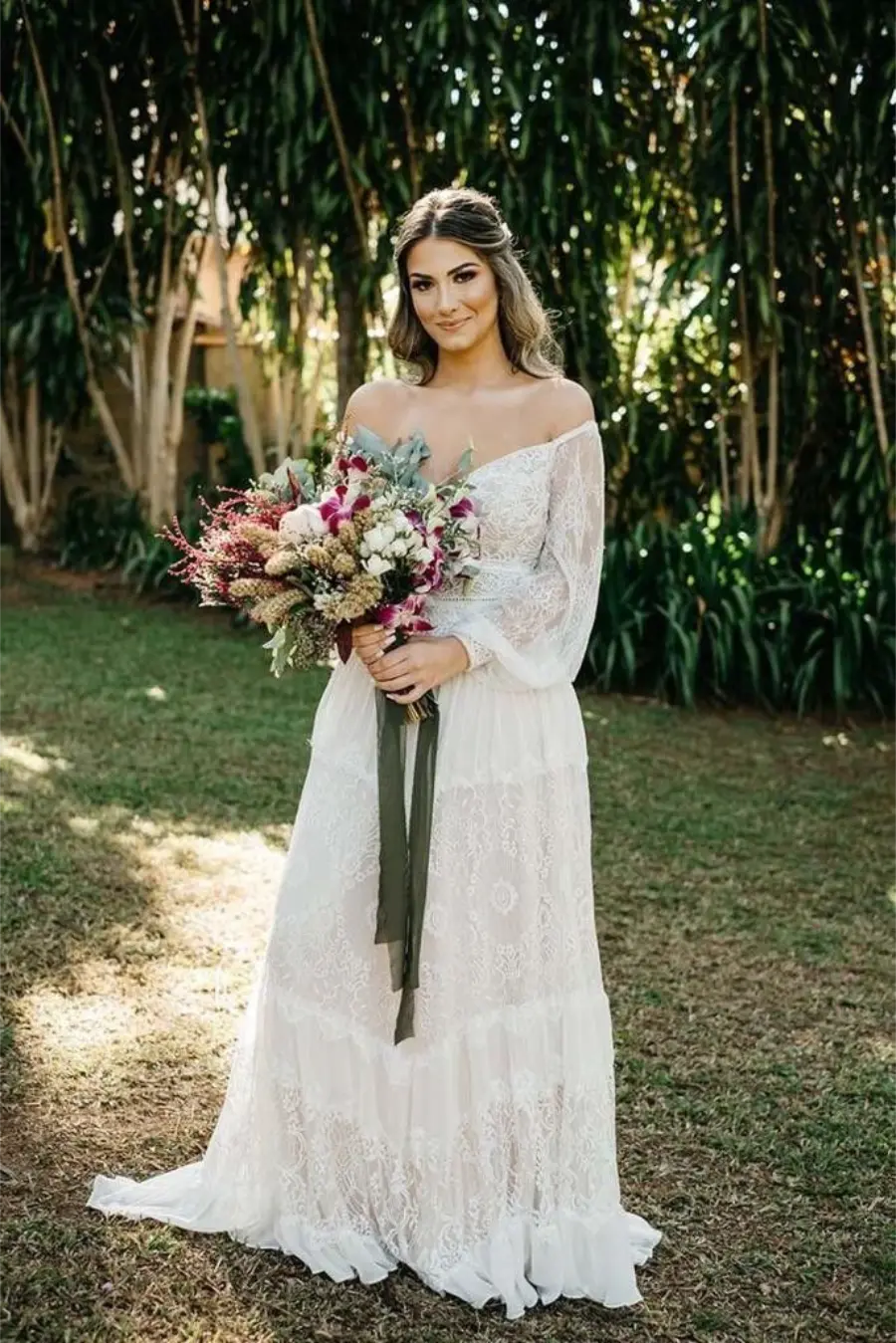 Noiva em vestido de renda ombro a ombro, segurando um buquê colorido com flores secas e naturais, em ambiente externo verde.