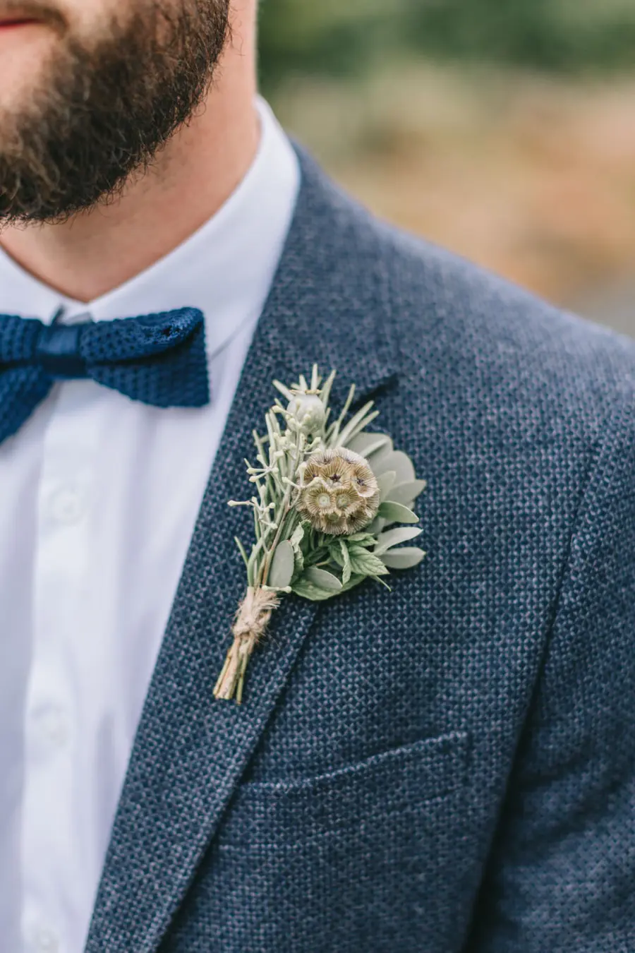 Detalhe de padrinho com gravata borboleta azul em terno de linho, boutonni&egrave;re r&uacute;stico e camisa branca