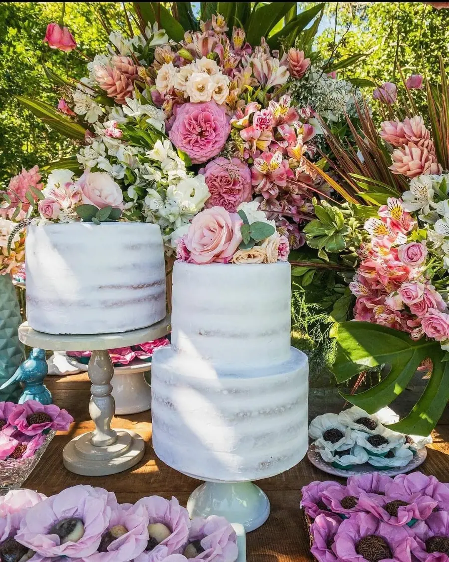 Conjunto de bolos brancos estilo semi naked decorados com rosas e folhagens, compondo uma mesa rústica com arranjo floral ao fundo.