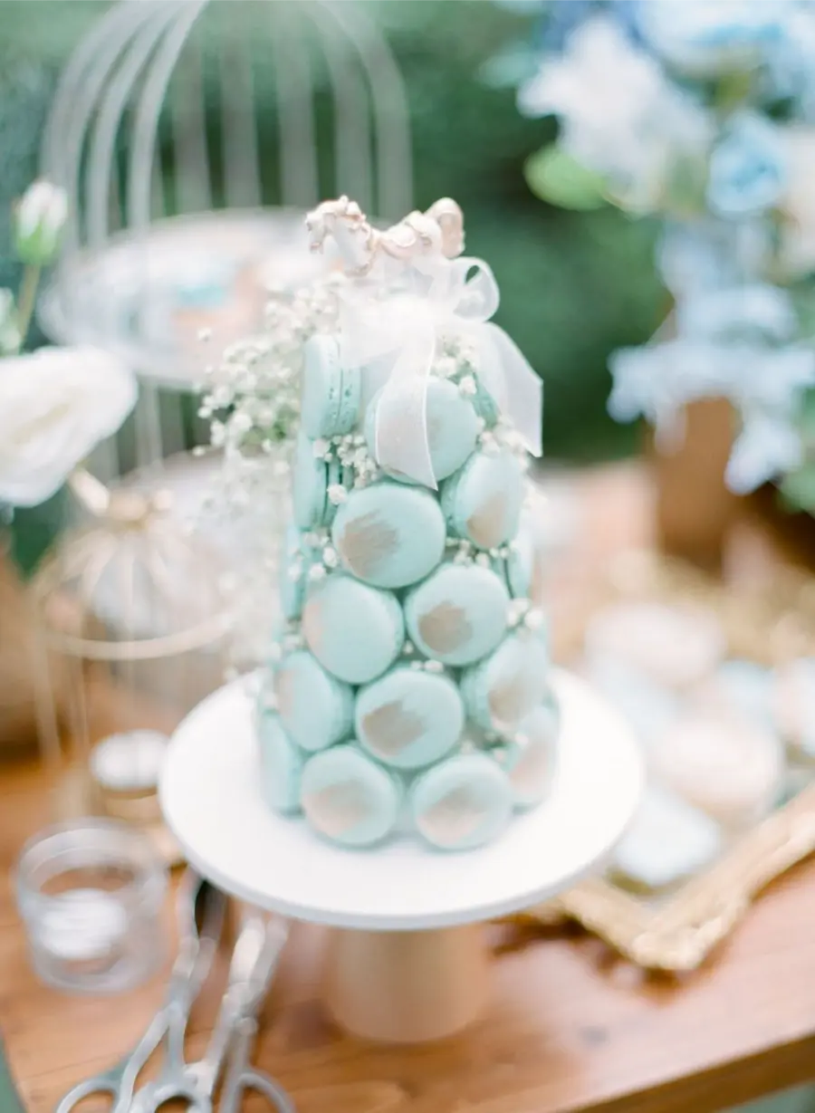 Torre de macarons azuis decorada com la&ccedil;o branco e flores delicadas, sobre base de bolo branca.