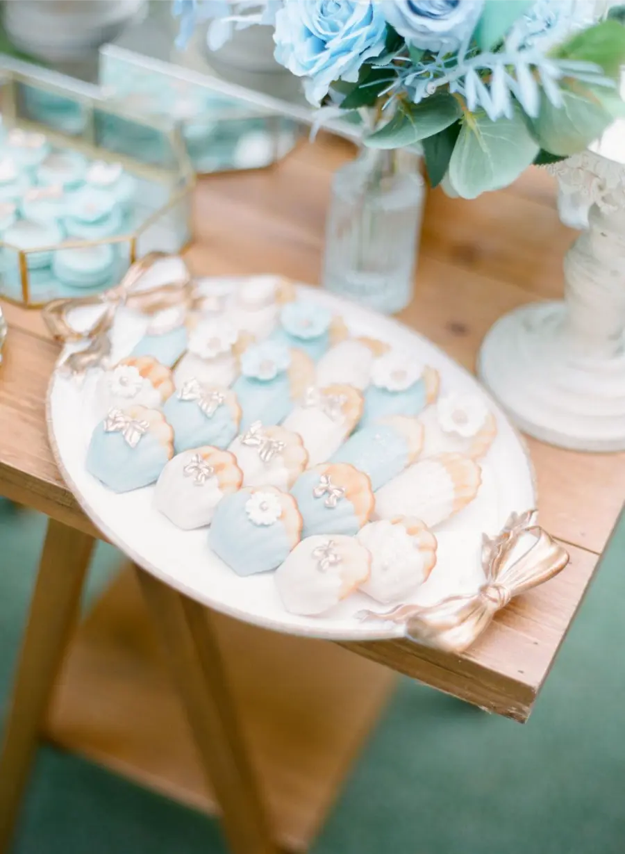 Biscoitos decorados em azul claro e branco, dispostos em prato branco com la&ccedil;os dourados nas laterais.