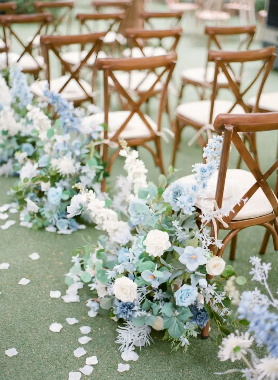 Decora&ccedil;&atilde;o de corredor de casamento com cadeiras de madeira e arranjos de flores em azul serenity, branco e folhagens verdes.