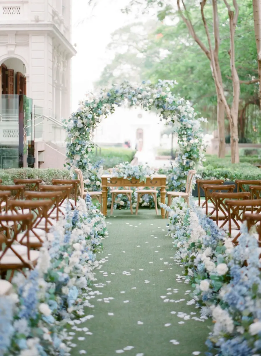 Cerim&ocirc;nia de casamento com arco e caminho decorados em flores azul serenity e brancas, criando ambiente rom&acirc;ntico ao ar livre.