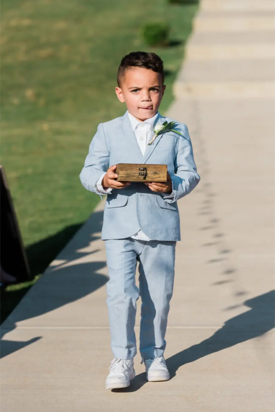 Menino pajem com terno azul serenity carregando caixinha de alian&ccedil;as durante entrada.