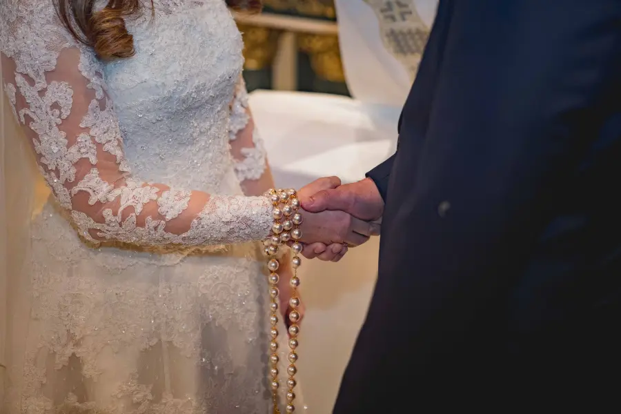 Noiva e noivo dando as mãos durante cerimônia com terço de pérolas douradas.