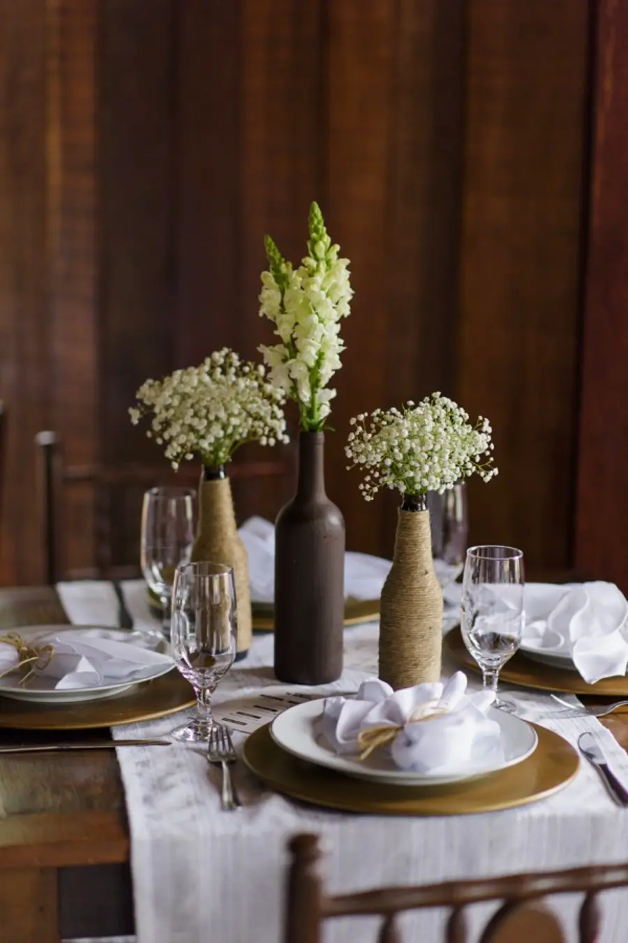 Mesa de recepção decorada com garrafas cobertas de corda e flores brancas, acompanhada de pratos e taças