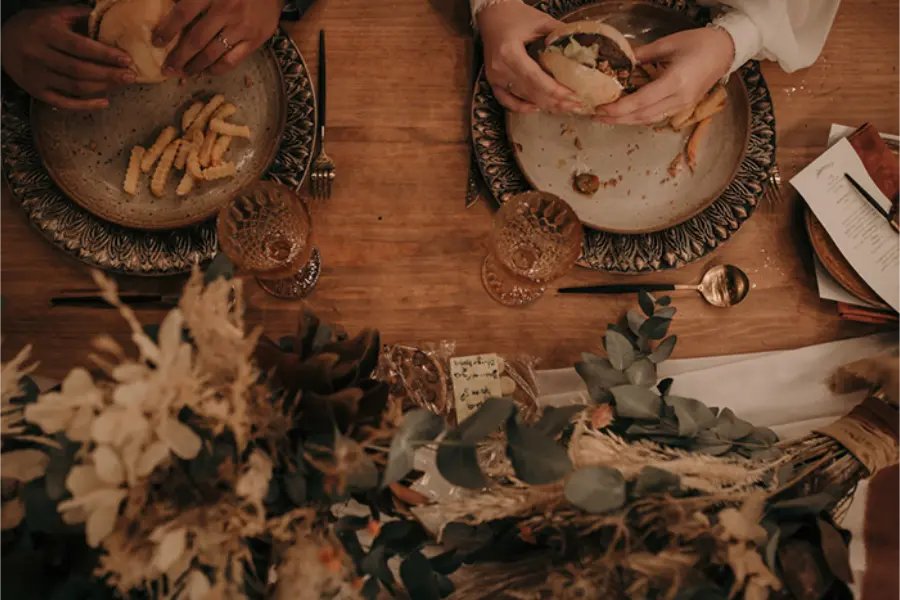 Casal de noivos compartilhando hambúrguer com batata frita durante a recepção.