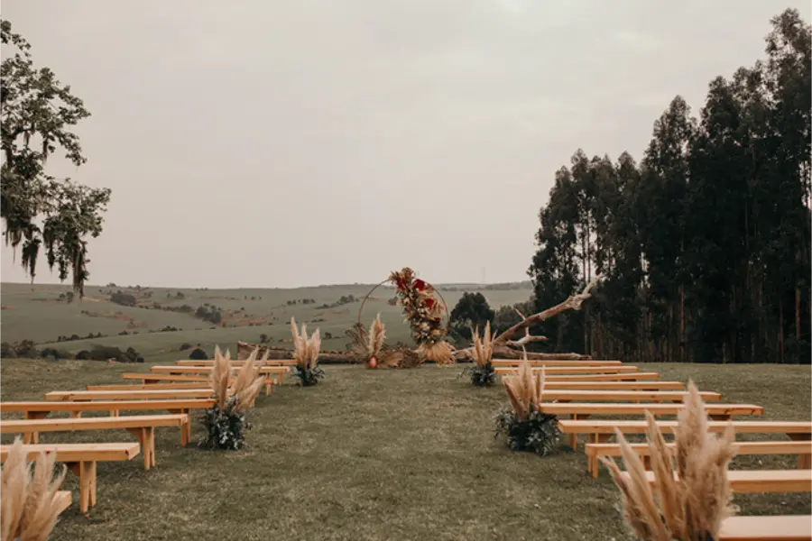 Banco de madeira alinhados em gramado e altar com arranjo de flores secas em espaço aberto no campo para cerimônia de casamento.