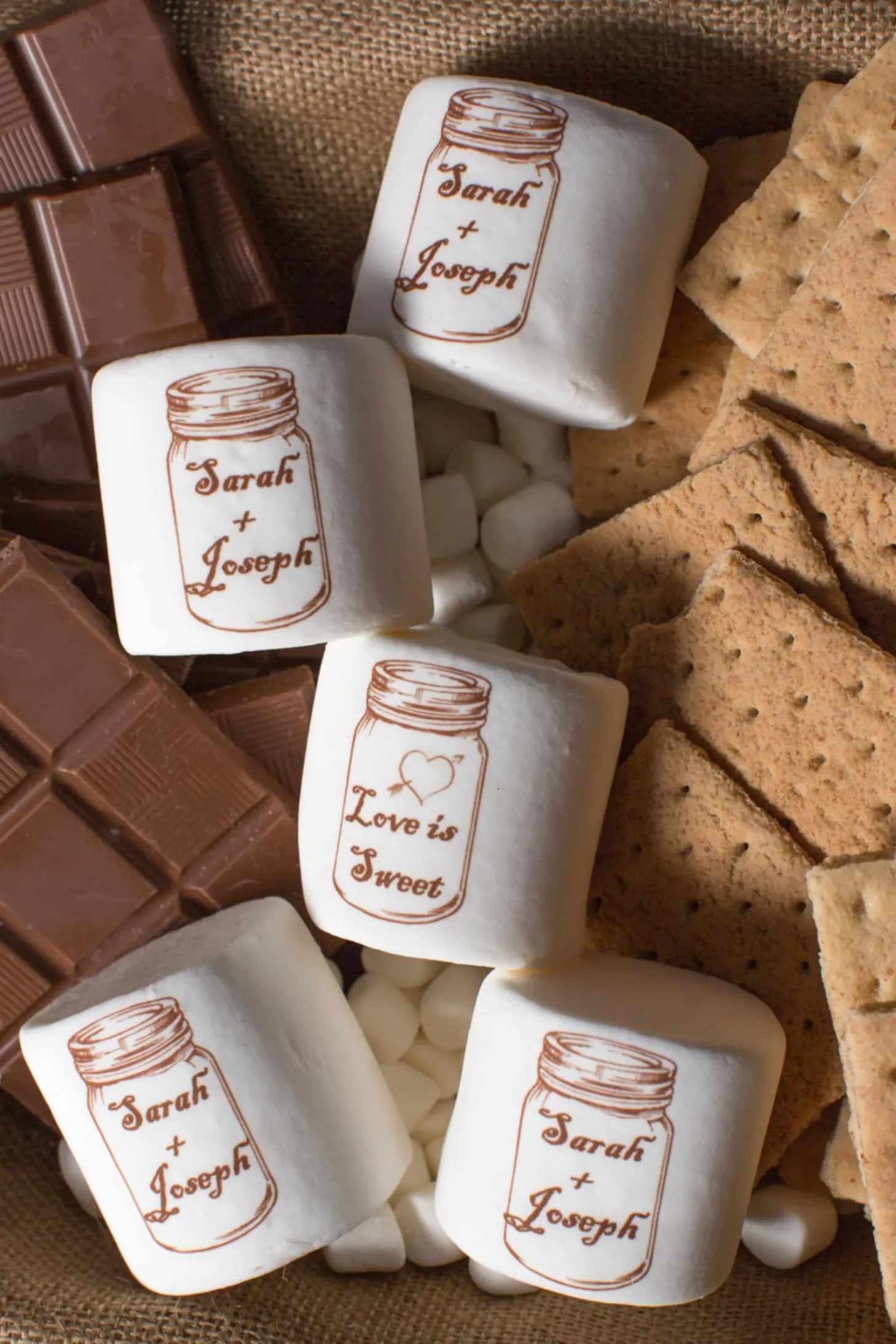 Marshmallows personalizados com nomes dos noivos e frases românticas, acompanhados de chocolate e biscoitos, lembrancinha criativa de casamento.