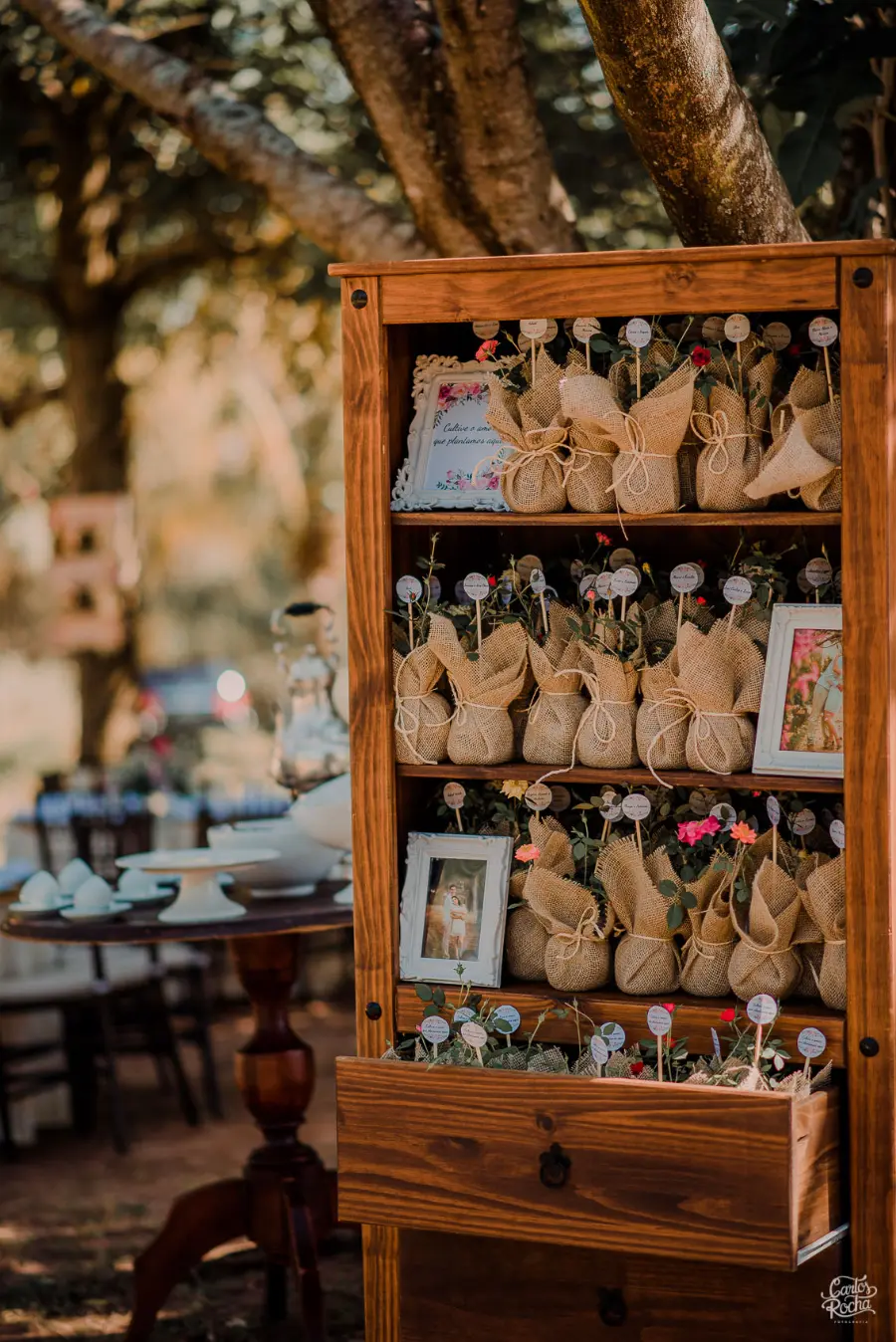 Prateleira de madeira repleta de lembrancinhas em juta com flores e plaquinhas decorativas, acompanhada de porta-retratos românticos.