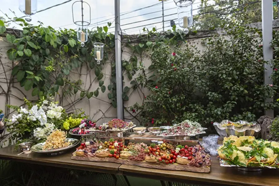 Buffet de casamento simples ao ar livre com massas, saladas e petiscos servidos em mesa de madeira com flores naturais.