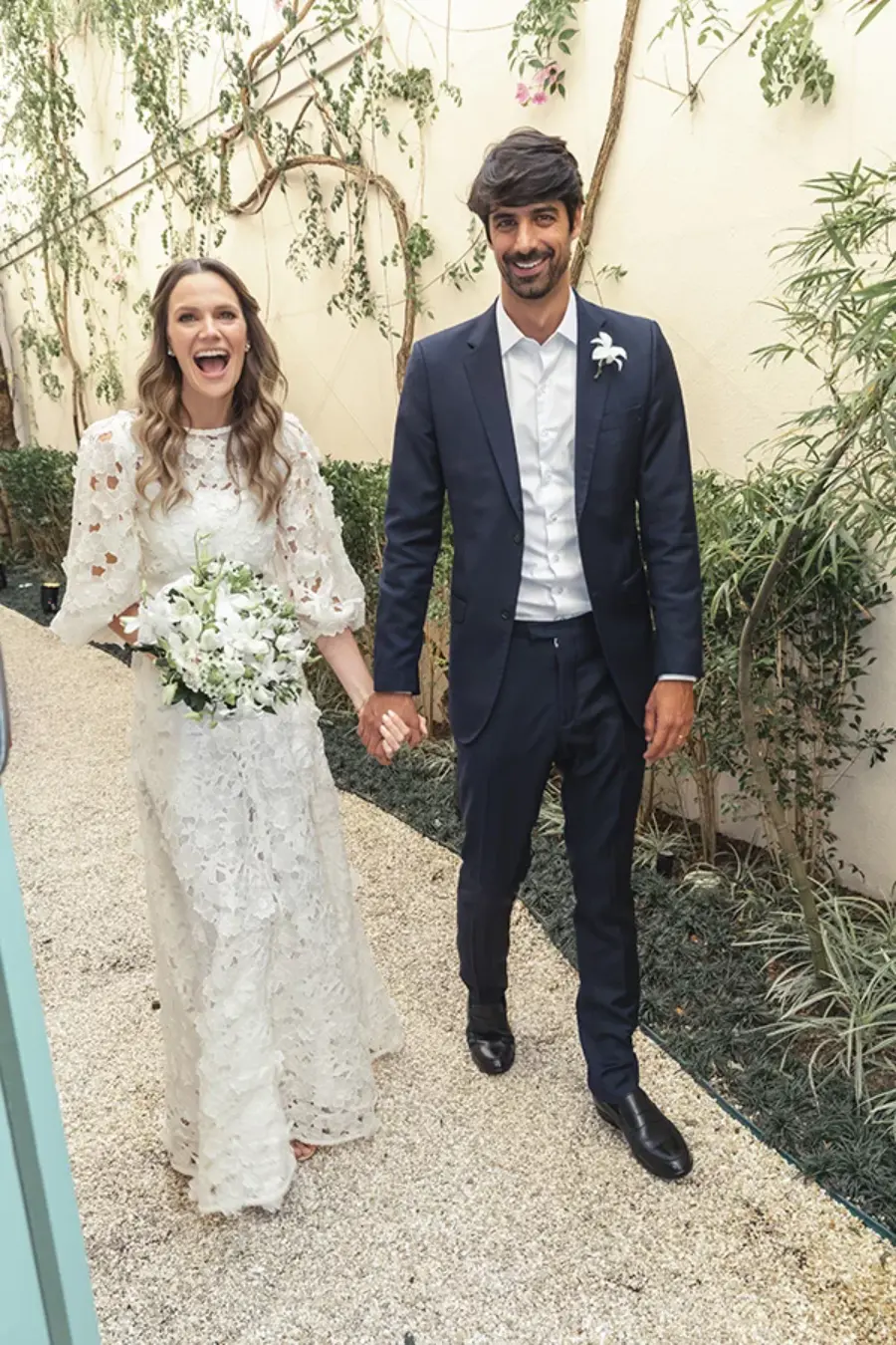 Noiva com vestido rendado e buquê de flores brancas caminha ao lado do noivo de terno azul, ambos sorrindo e de mãos dadas.
