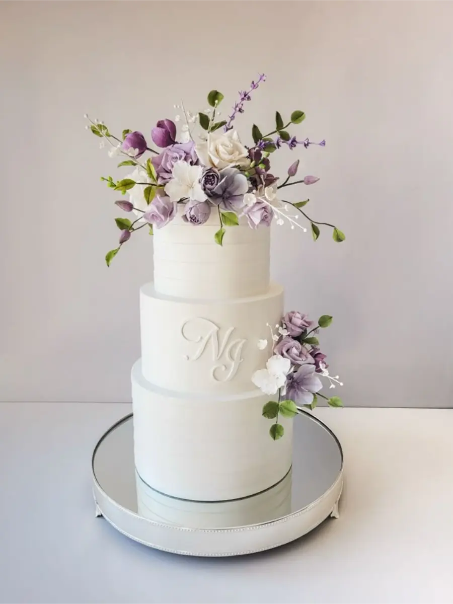 Bolo cenográfico de três andares branco com flores lilás e iniciais personalizadas NG